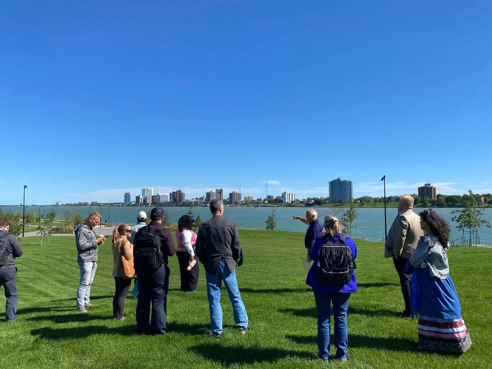 A group of people stand together on green grass overlooking the Detroit River.