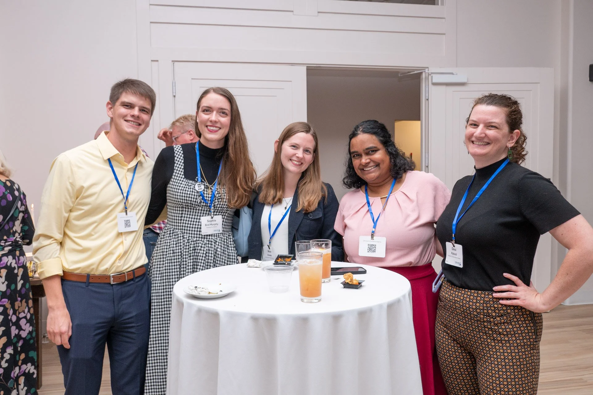alt="five young people stand together smiling around a small circular table covered with white linen."