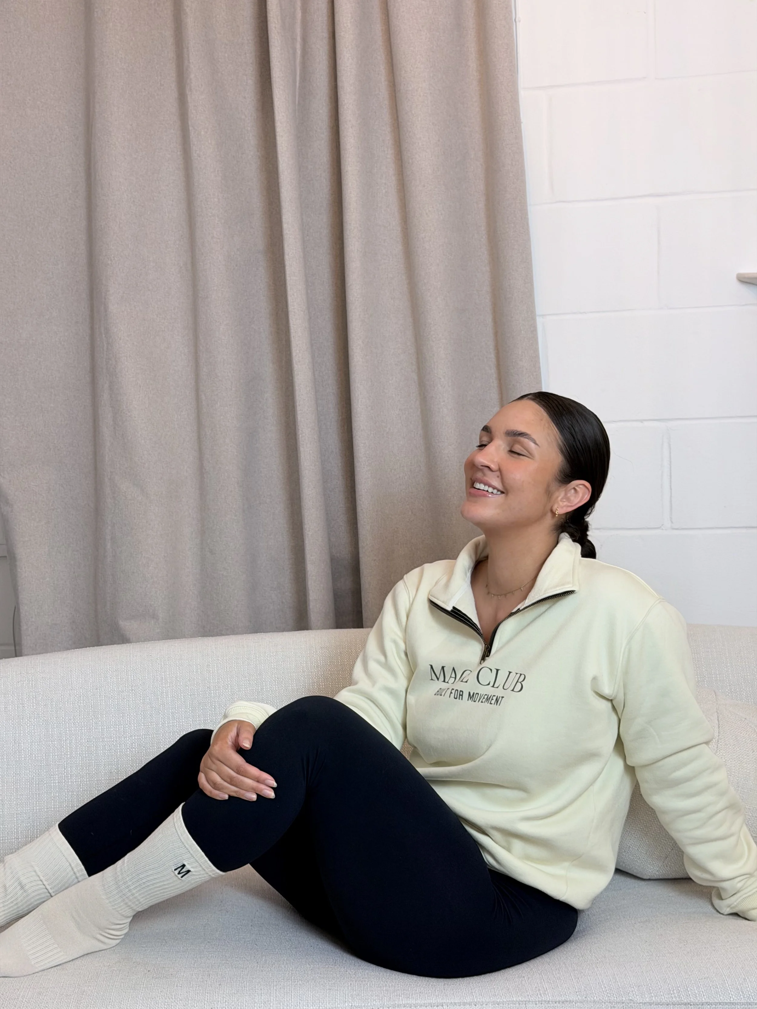 A woman sitting on a sofa, smiling with eyes closed, wearing a cream-colored zip-up sweatshirt, black leggings, and white socks, in a room with beige curtains and white brick wall.