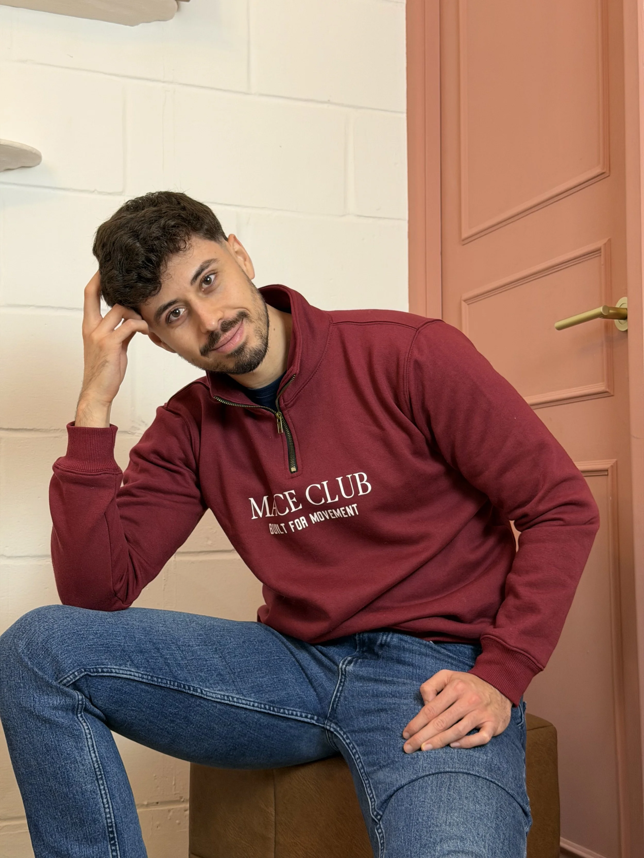 A young man with dark hair and a beard, wearing a maroon zip-up sweatshirt that says MACE CLUB BUILT FOR MOVEMENT, sitting on a brown ottoman, leaning slightly to the left, with his right hand resting on his head and his left hand on his knee, in front of a white wall and a pink door.