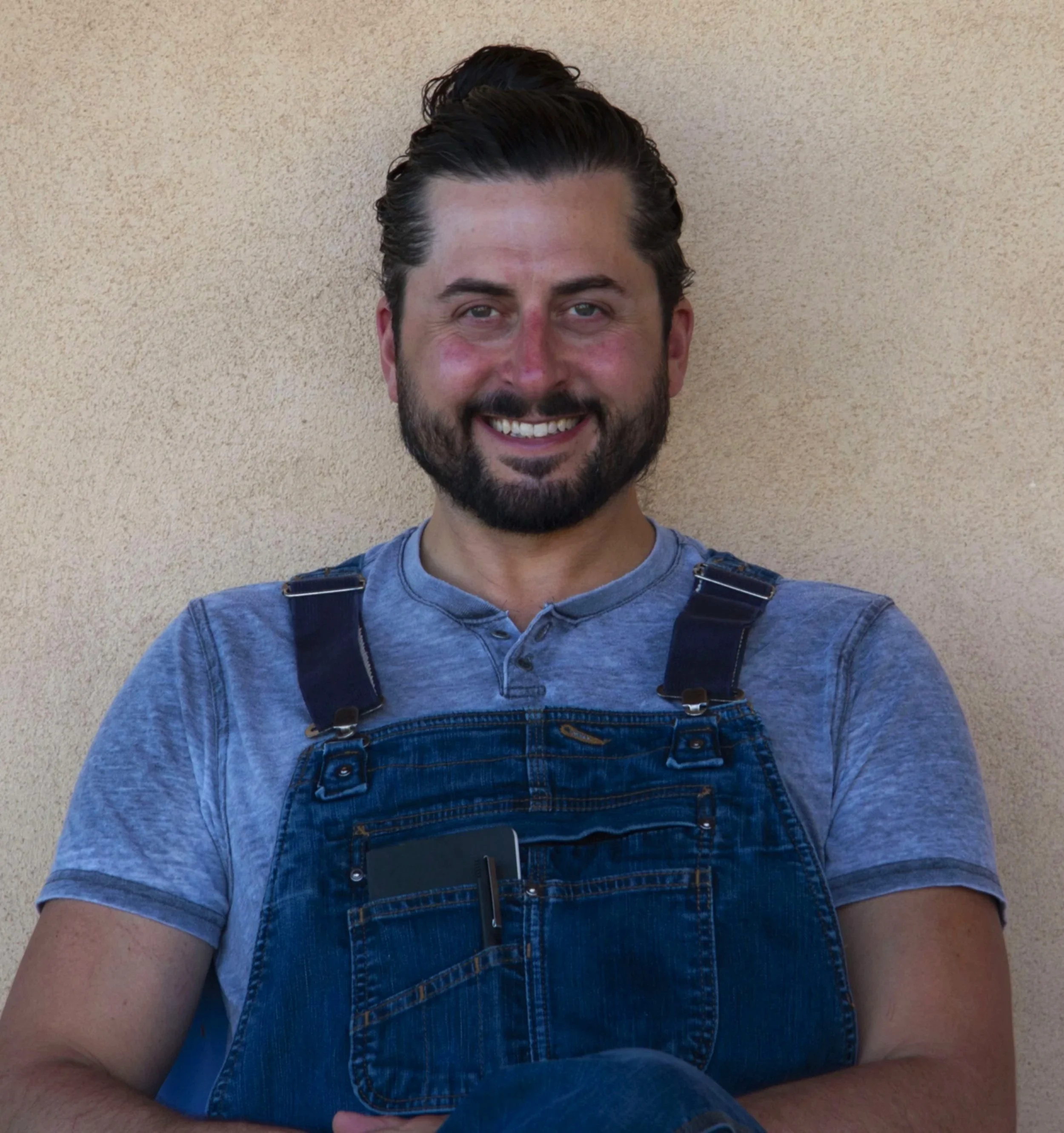 A smiling man with a beard and mustache, wearing a gray T-shirt and denim overalls, photographed against a beige wall.