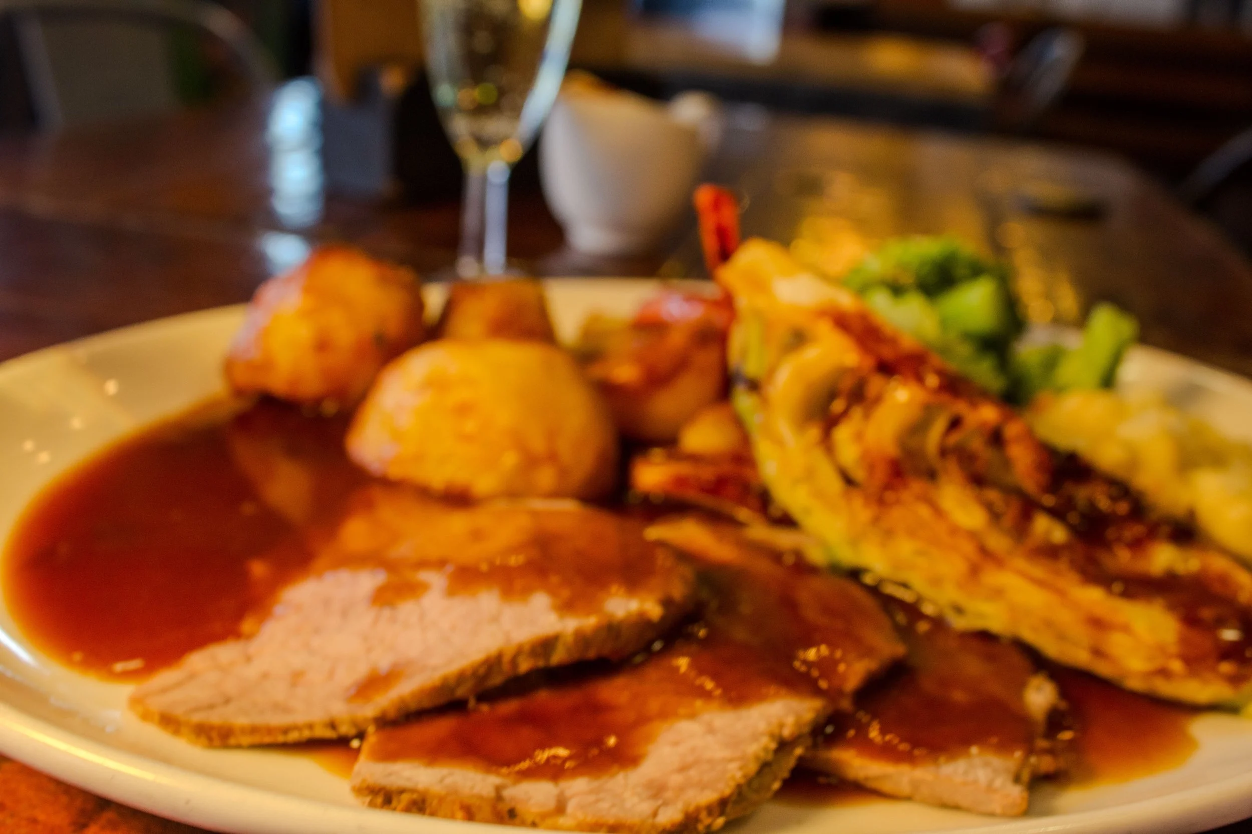 A plate of food with roasted potatoes, grilled chicken, vegetables, and a dark sauce, with a glass of wine and a bowl in the background on a wooden table.