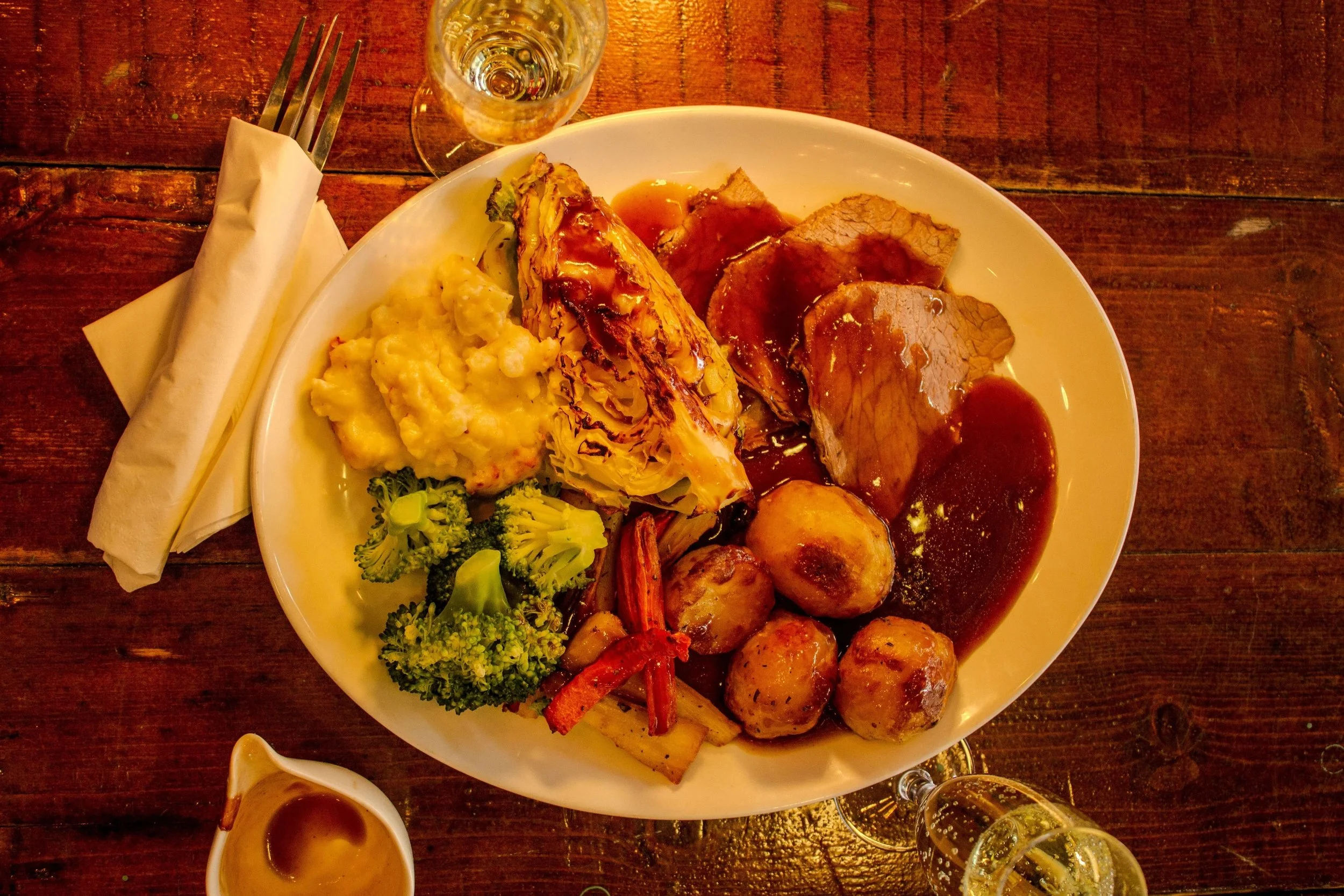 A plate of assorted food items including mashed potatoes, broccoli, roasted vegetables, and slices of meat with gravy, accompanied by a glass of white wine and a small dish of gravy on a wooden table.