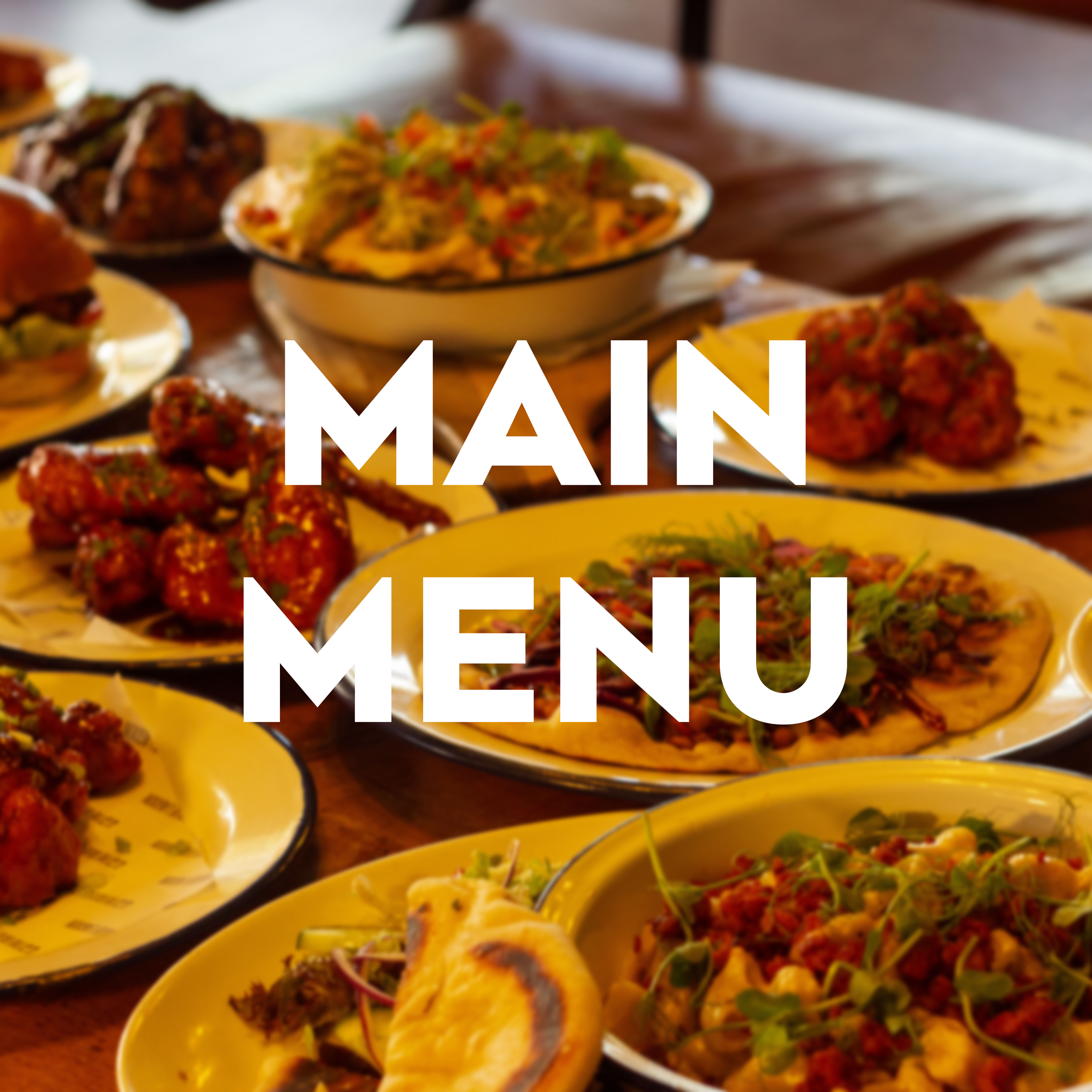 Main Menu text written over an image of a table with several plates of food, including flatbreads and salads, topped with various ingredients, and side dishes, set in a restaurant or dining area.