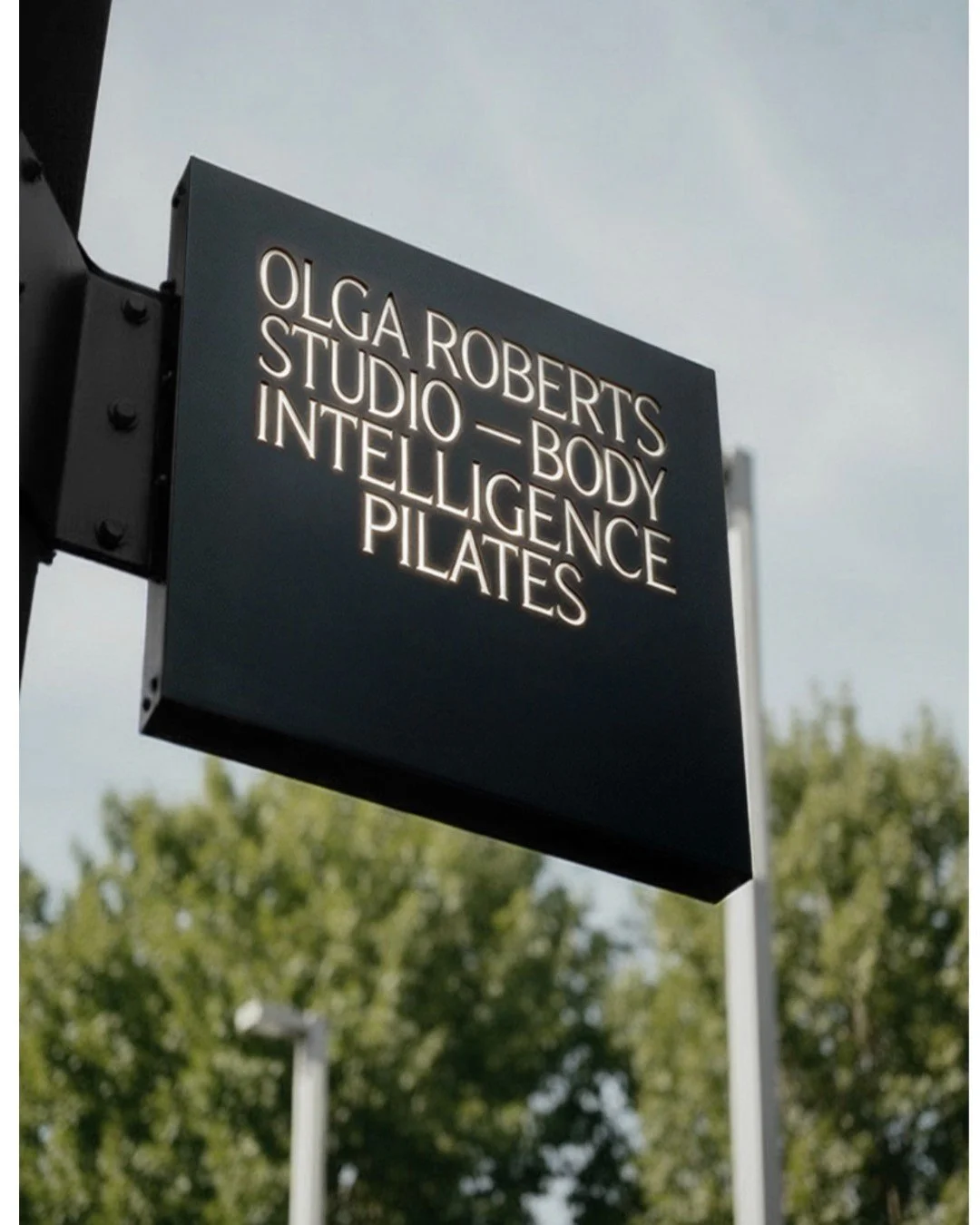 Black sign hanging outdoors with neon illuminated text that reads: Olga Roberts Studio - Body Intelligence Pilates, against a background of green trees and a cloudy sky.