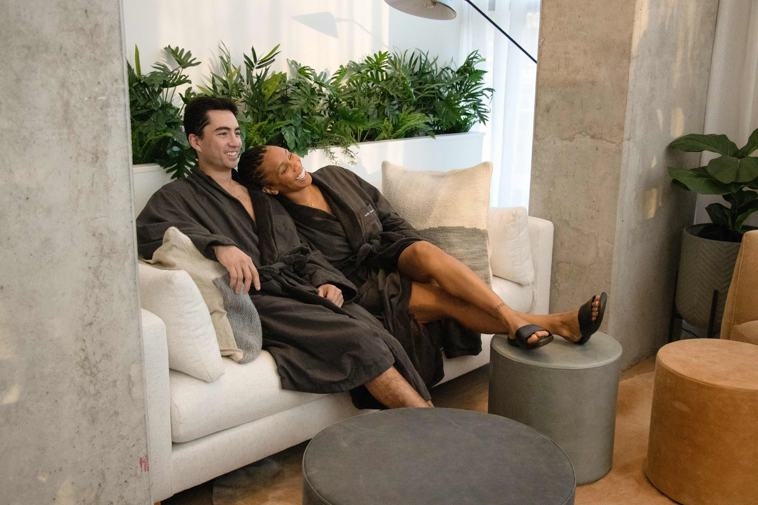 Two people relaxing on a white sofa with cushions in a modern, green indoor space. They are wearing black robes and smiling, with one person's leg resting on a round ottoman.