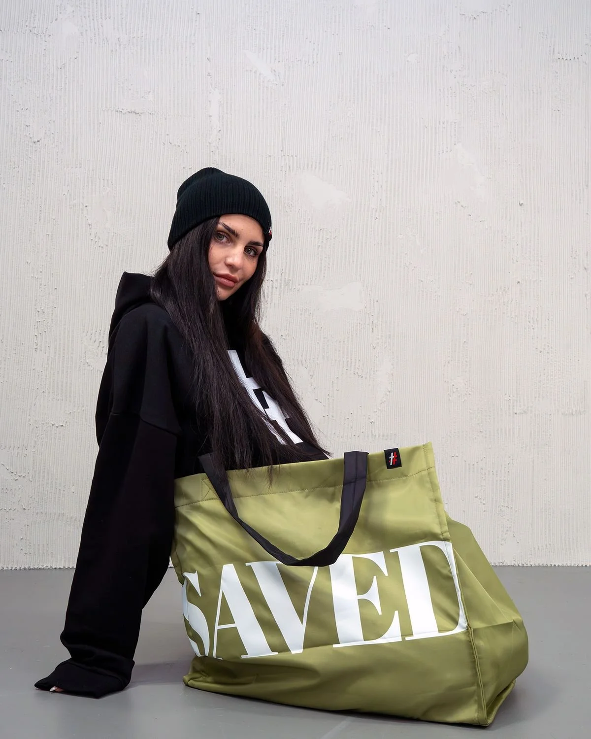 Woman in a black beanie leaning on an olive SAVED bag