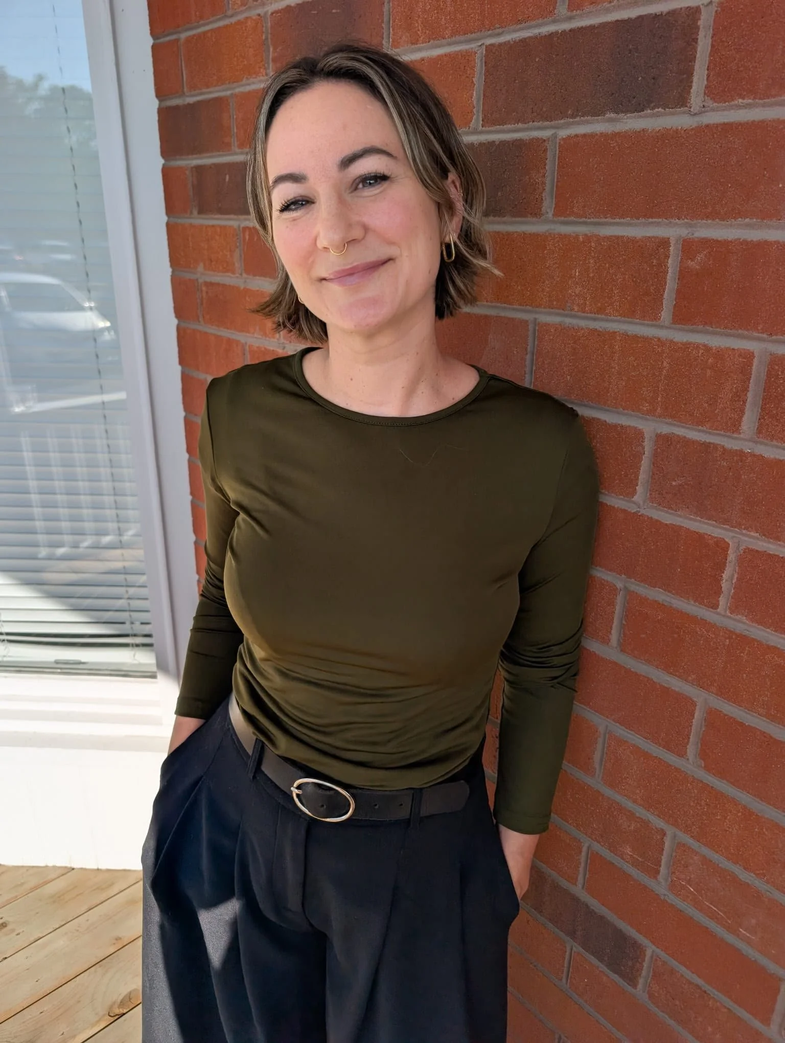 A person with short hair and a bright smile, wearing a sleeveless black top and patterned pants, stands against a red brick wall.