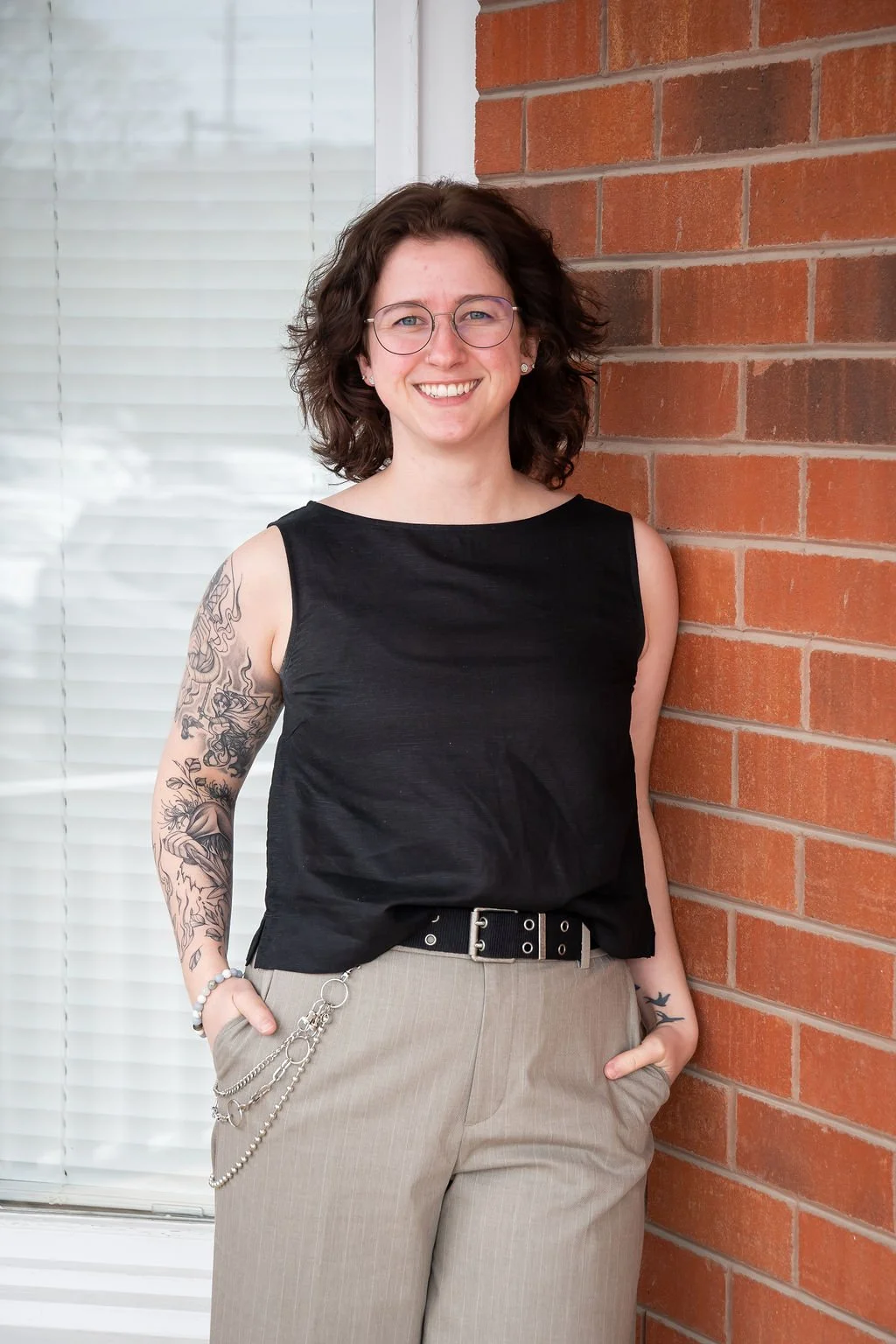 A smiling woman with curly reddish hair and glasses, wearing a white blouse and black pants, standing against a brick wall.