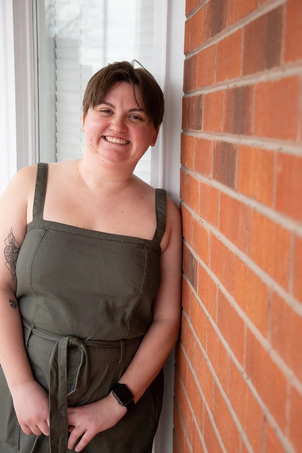 A young person with short brown hair smiling and standing against a brick wall, wearing a black top, striped shirt, light blue jeans, and a black belt, with sunlight casting shadows.