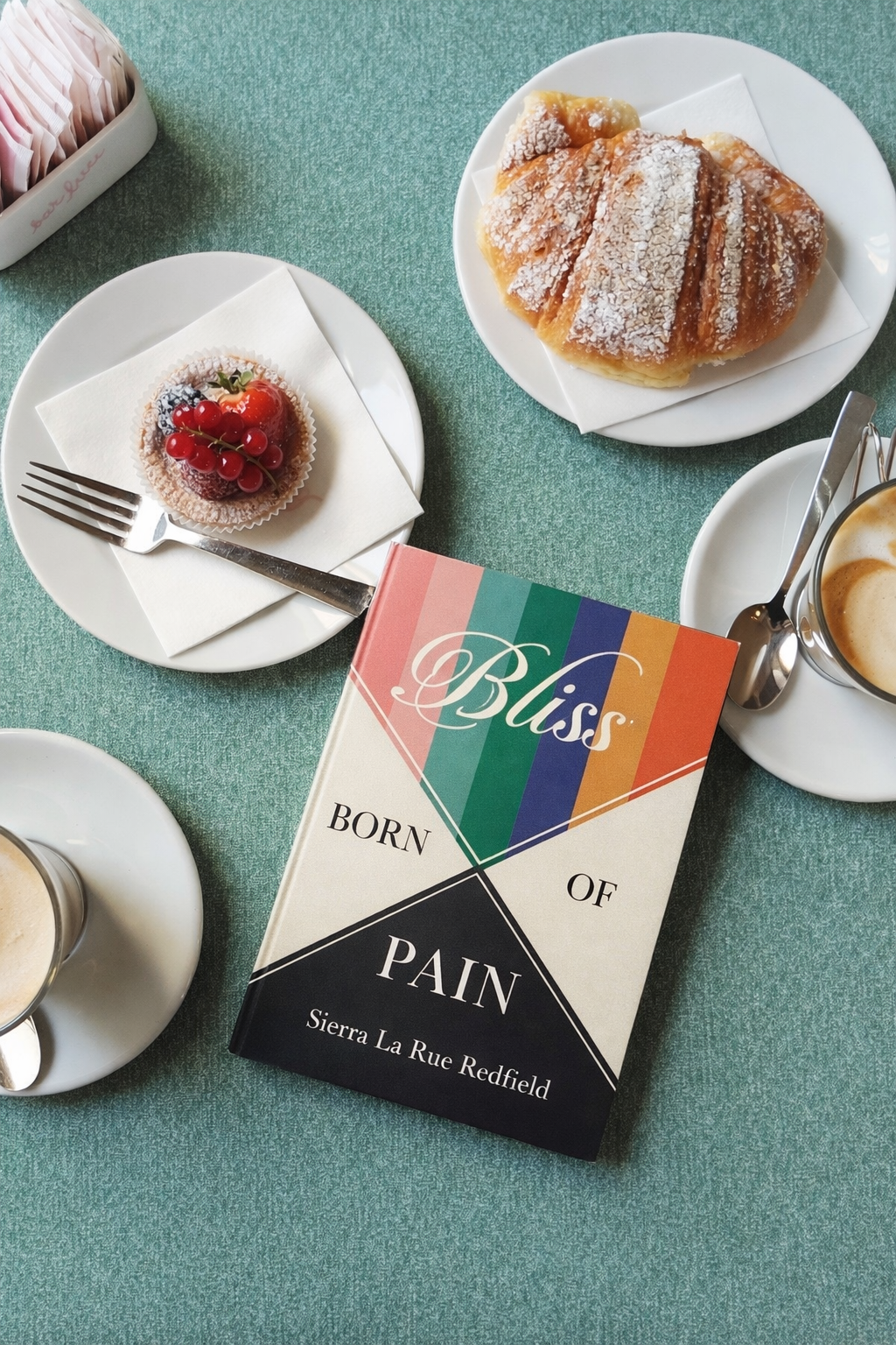 Bliss Born of Pain by Sierra La Rue Redfield on a café table with coffee and pastries.
