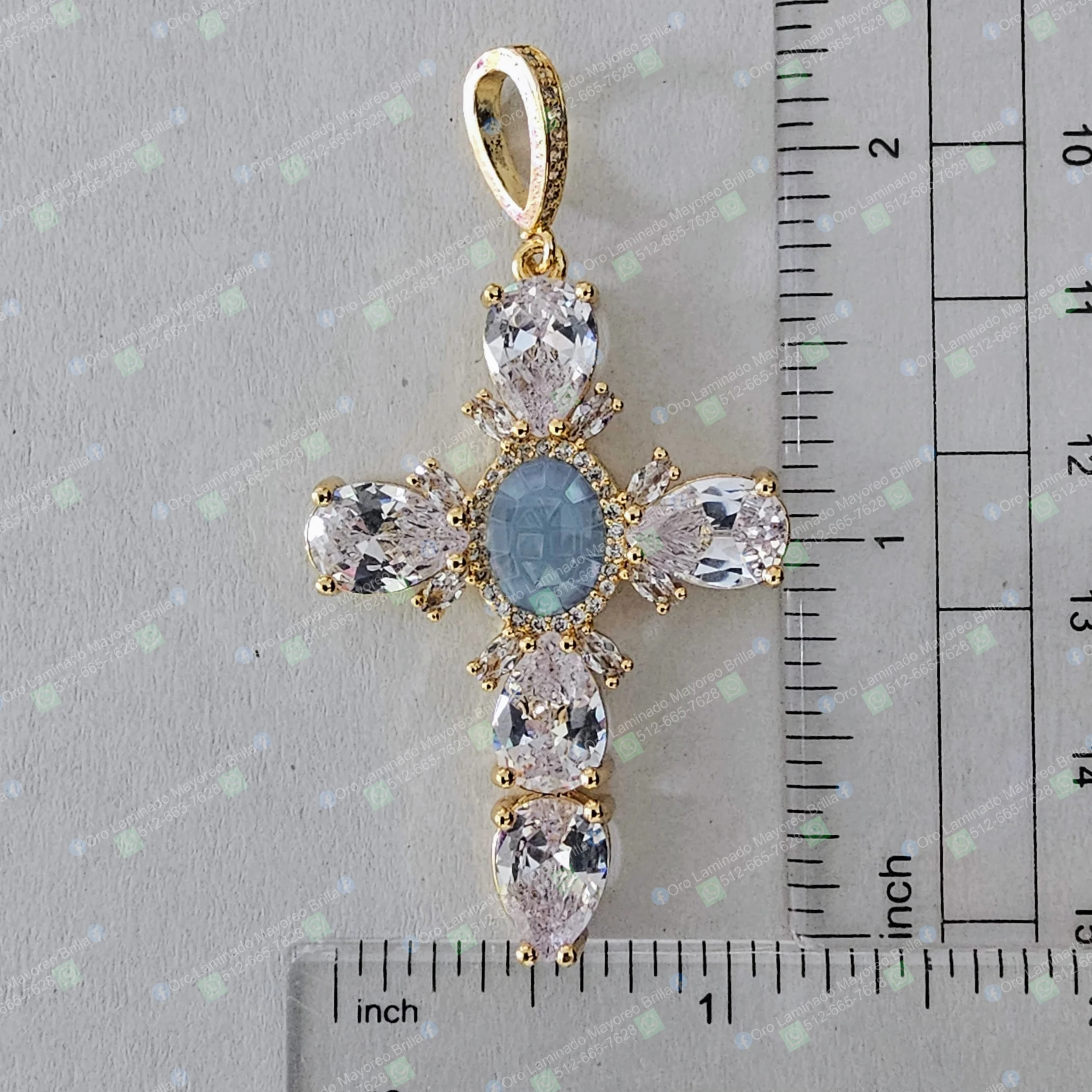 Dije cruz color oro con piedra azul central y cristales decorativos visibles | Gold color cross pendant with central blue stone and visible decorative crystals
