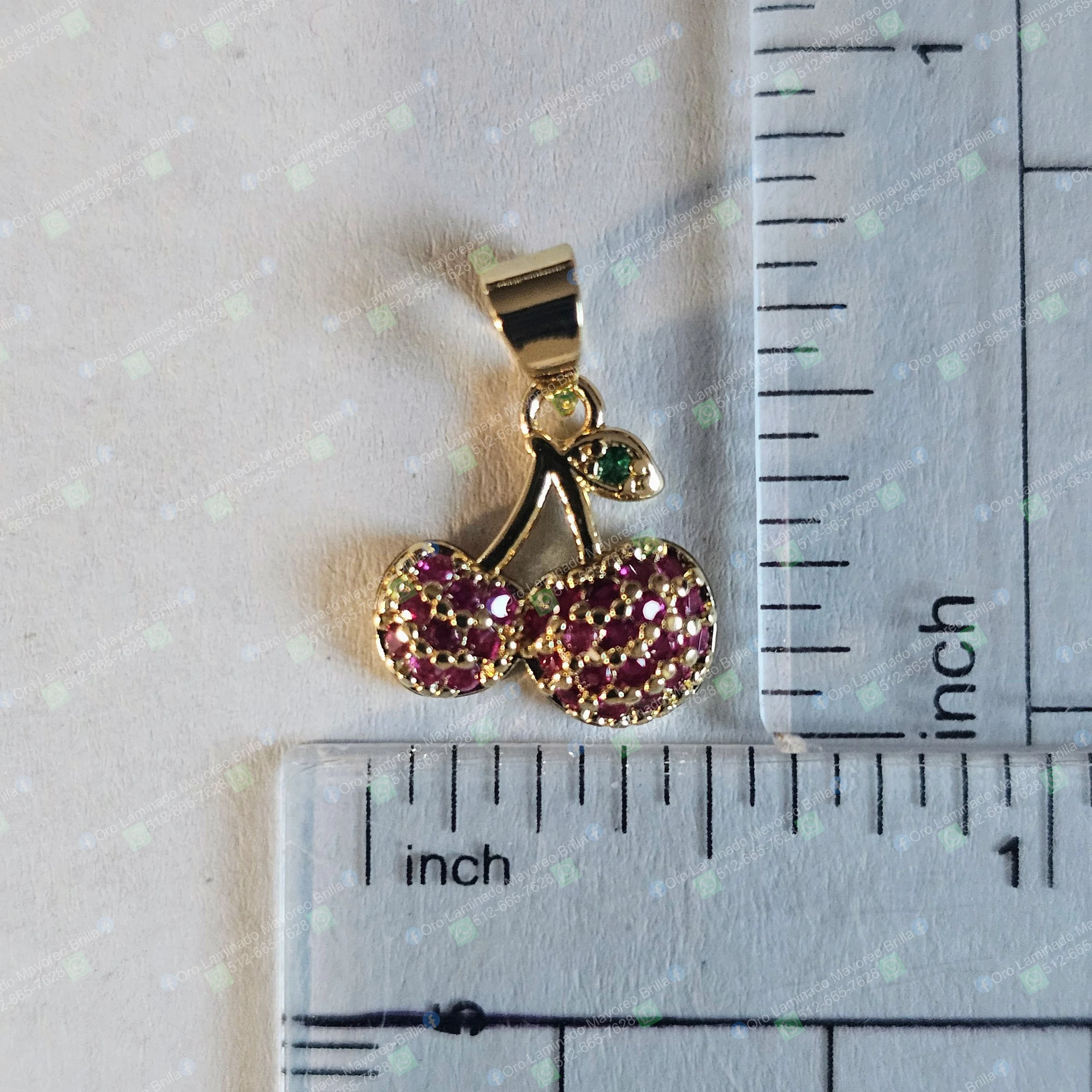 Dije de cereza color oro con piedras rojas decorativas y hoja con piedra verde visible | Gold color cherry pendant with red decorative stones and leaf with visible green stone