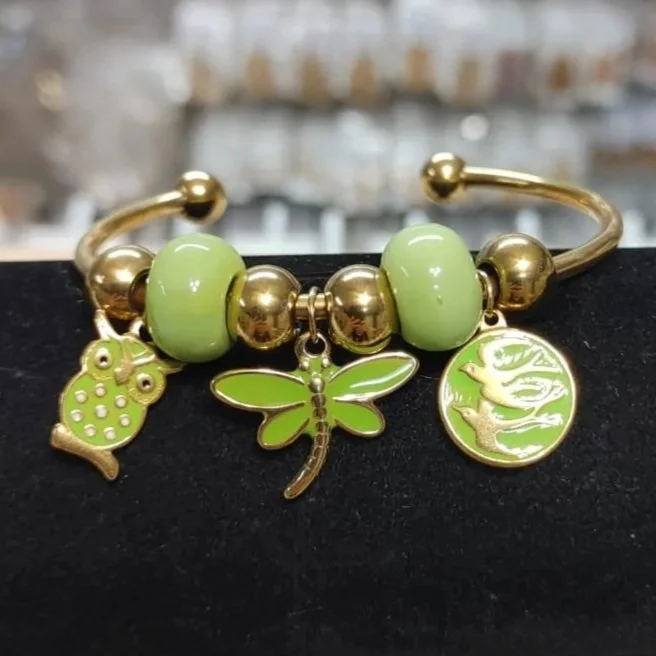 Pulsera dorada con dijes verdes de búho, libélula y ave en acero inoxidable. Gold bracelet with green owl, dragonfly, and bird charms in stainless steel.
