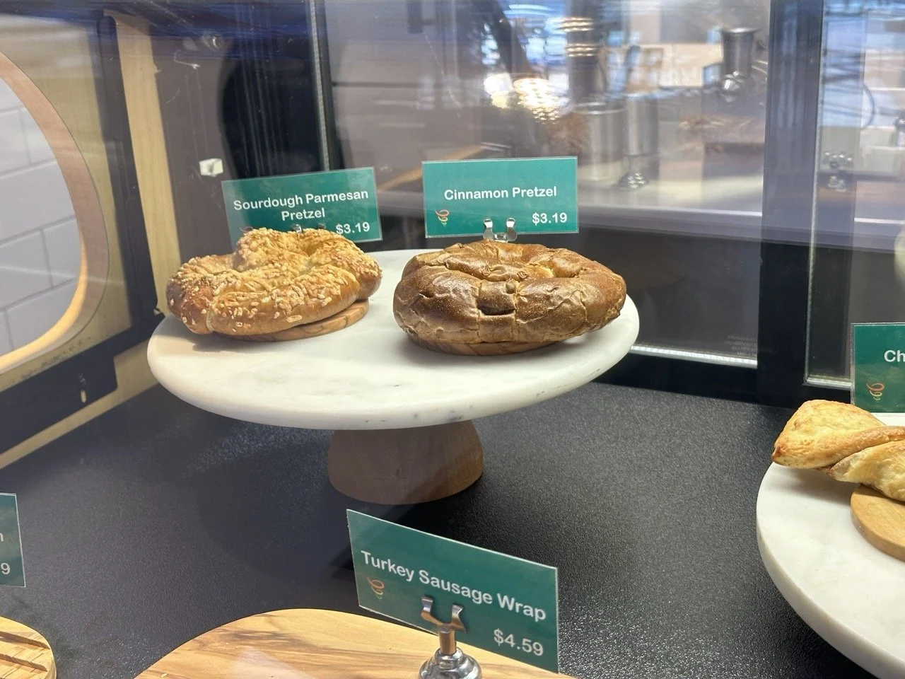 Display case with two pretzels, one sourdough parmesan and one cinnamon, priced at $3.19 each. A turkey sausage wrap priced at $4.59 is also visible.