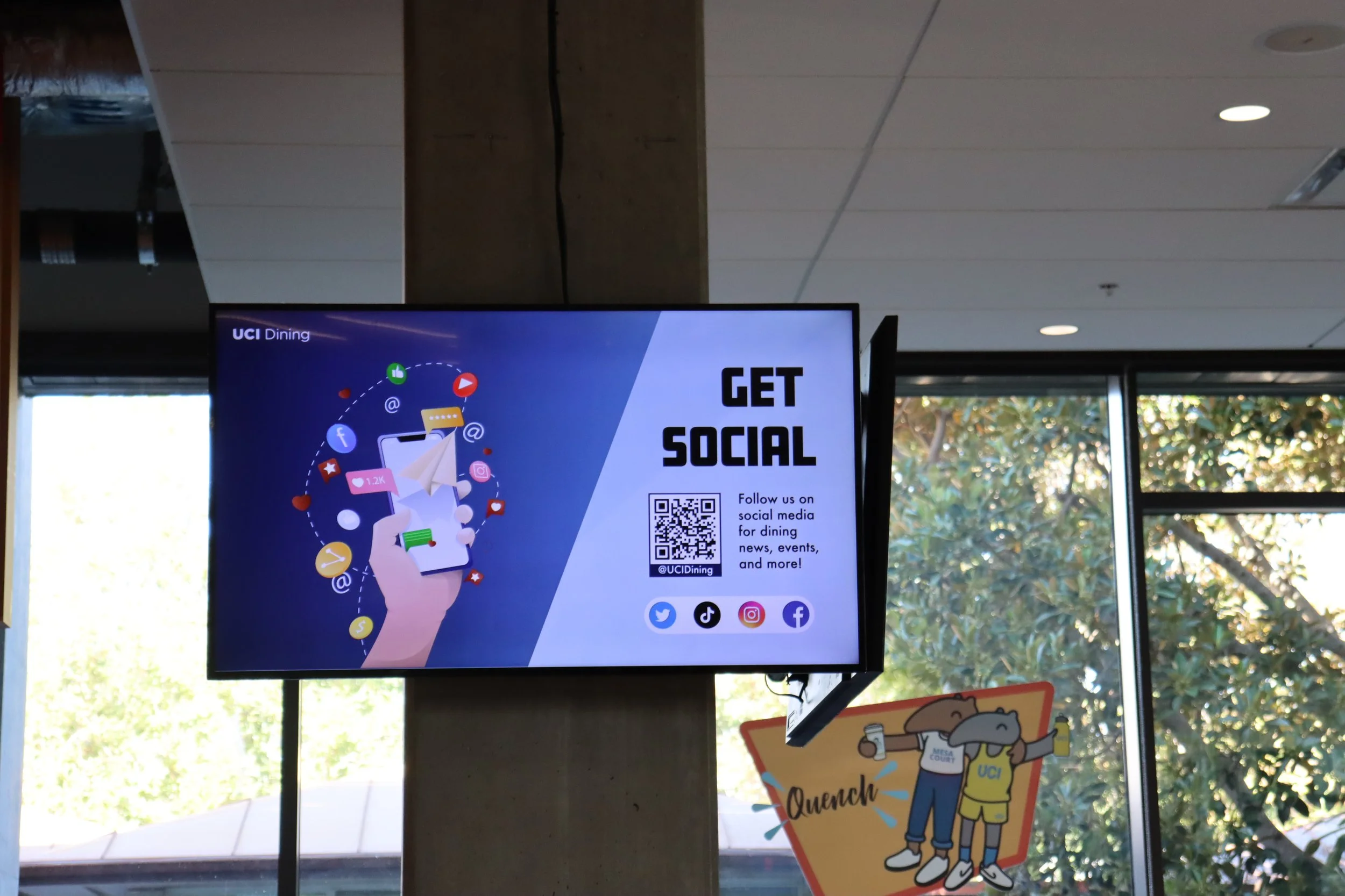 Digital sign promoting getting social on social media, featuring a QR code and icons for Twitter, TikTok, Instagram, and Facebook, with a background illustration of a hand holding a smartphone with app icons and messages.