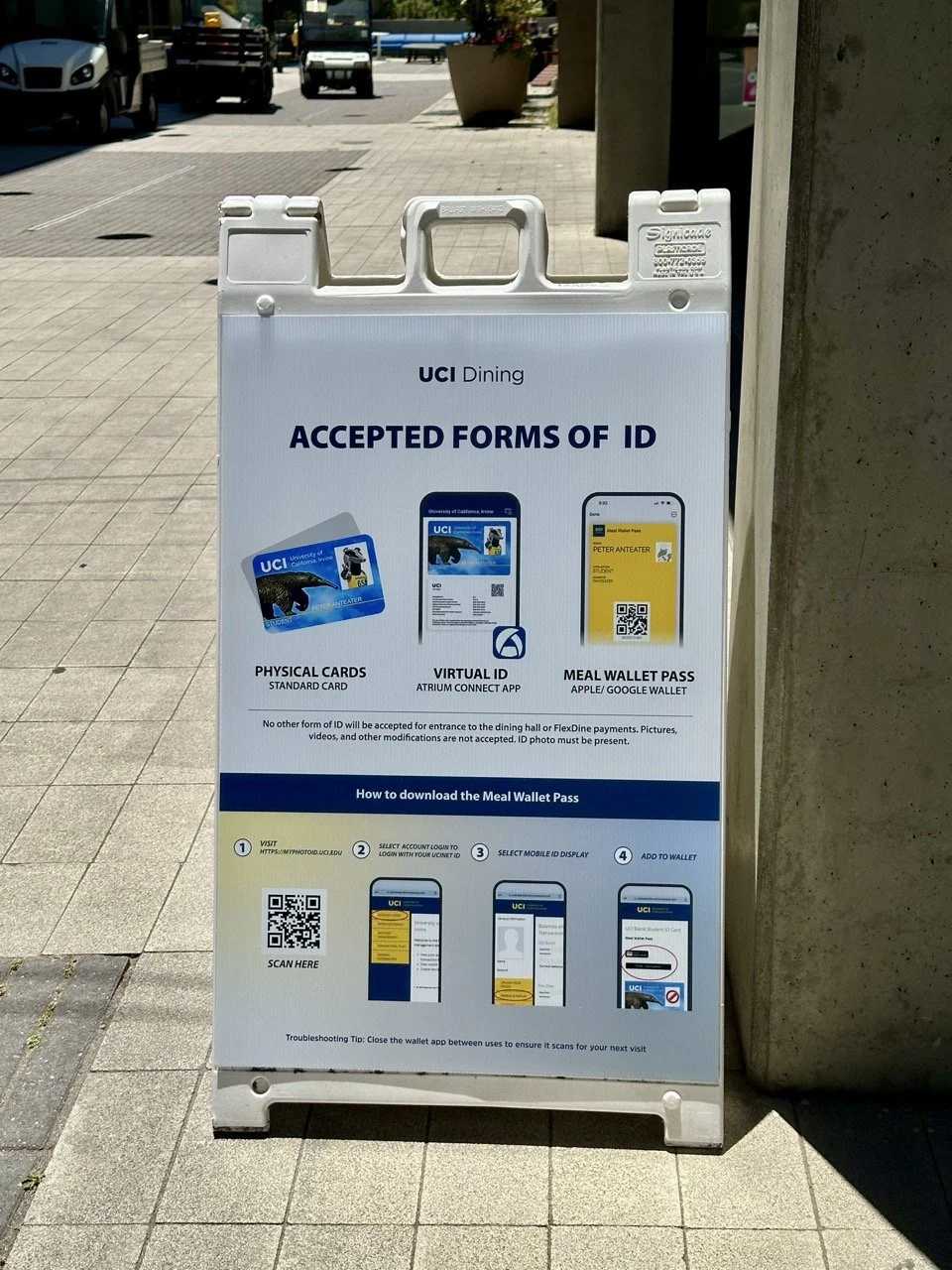 Sign displaying acceptable forms of ID for UCI Dining, including physical cards, virtual ID, and meal wallet pass, with instructions on how to download the meal wallet pass via QR code.