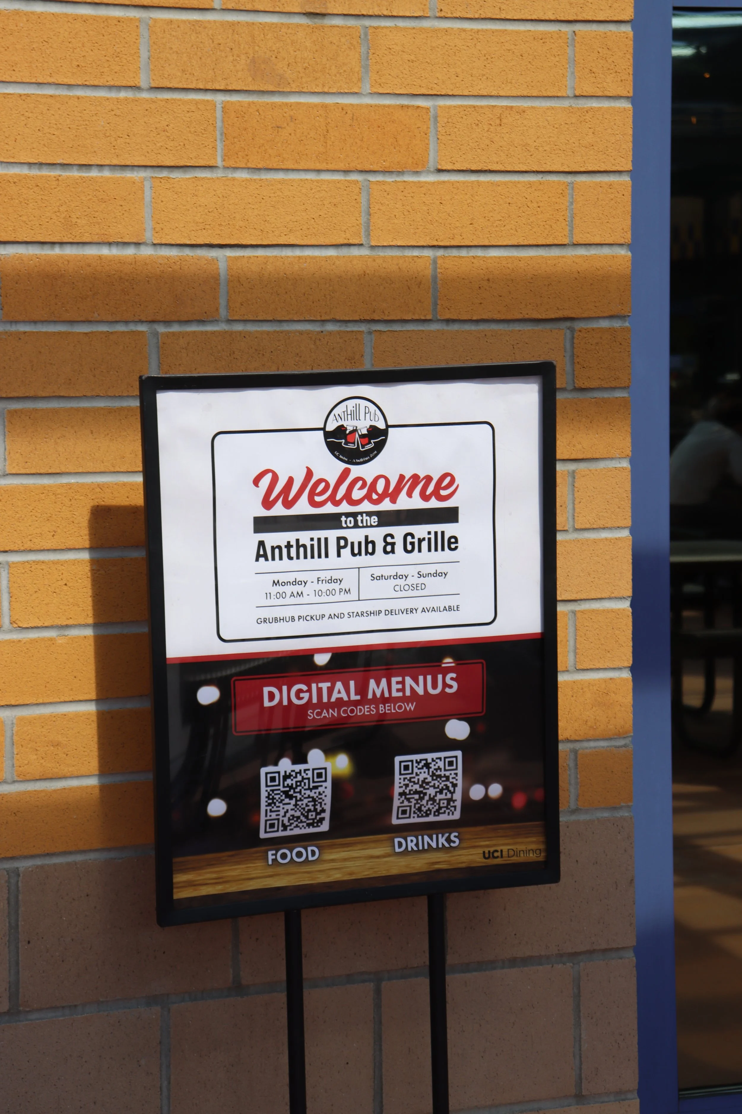 Signboard outside Anthill Pub & Grille displaying hours, digital menu QR codes for food and drinks, and opening days, mounted on a yellow brick wall.