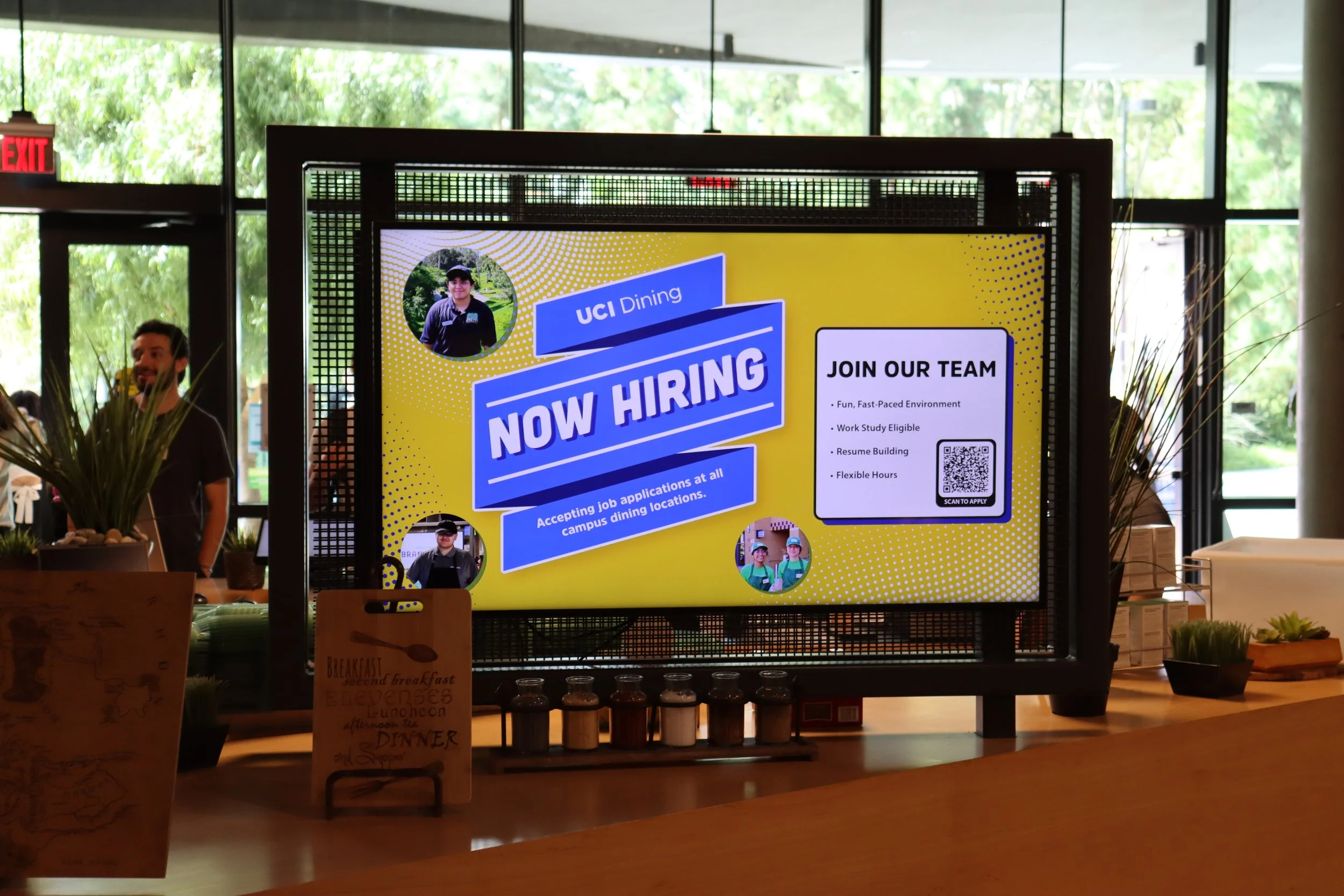 Digital sign in a campus dining area announcing job openings at UC Irvine Dining, with information about applying and a QR code. The background shows people and greenery outside through large windows.