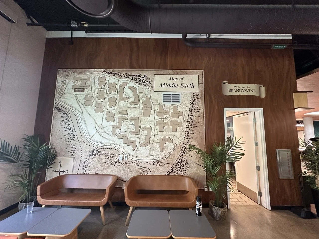 Map of Middle Earth mounted on a wooden wall in the lobby of Brandywine. Two brown chairs and a table with a potted plant and a cup are in front, next to an open door.