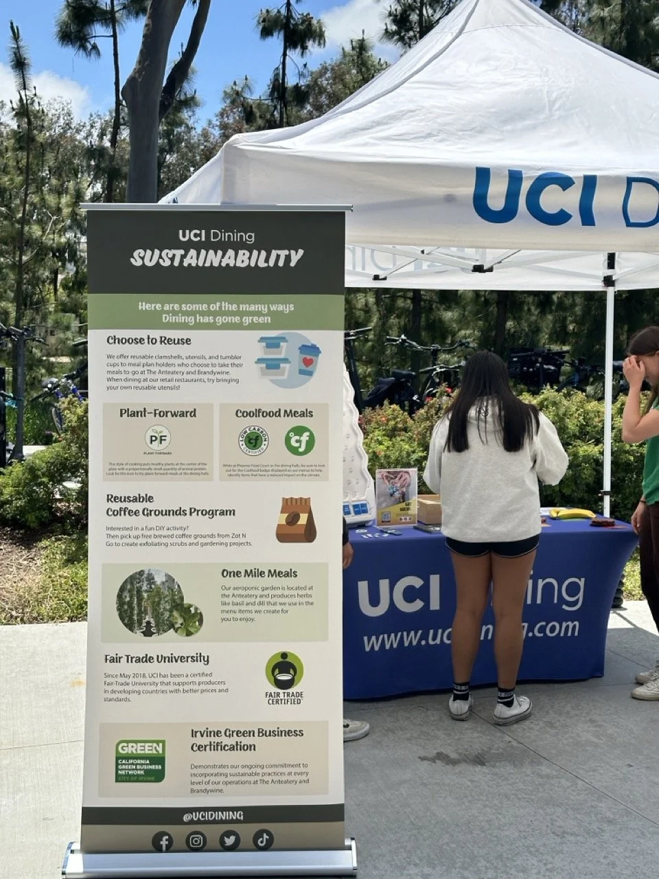 UCI Dining sustainability informational booth outdoors under a white canopy with a banner, with three people nearby and several bicycles in the background. There is a large vertical sign with tips for sustainable dining, including options like reusin