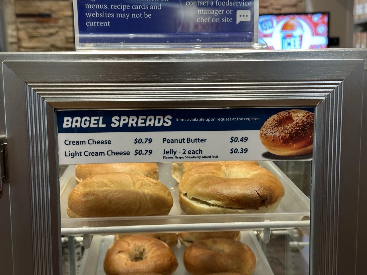 Display case with a variety of bagel spreads and bagels inside. The sign labels the spreads including cream cheese, light cream cheese, peanut butter, and jelly. The bagels are on a metal shelf, and a printed image of a bagel with sesame seeds is als