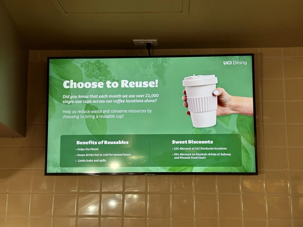 Digital sign encouraging reusable cups in UCI Dining, with a visual of a hand holding a white reusable coffee cup and text promoting benefits and discounts.