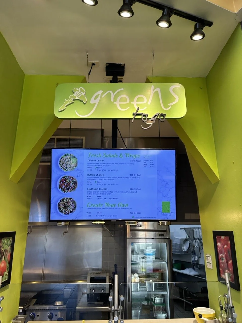 Green sign with white and black lettering for a restaurant called Greens to Go, menu screen displaying salads and wraps, in a bright yellow and green interior with kitchen appliances.
