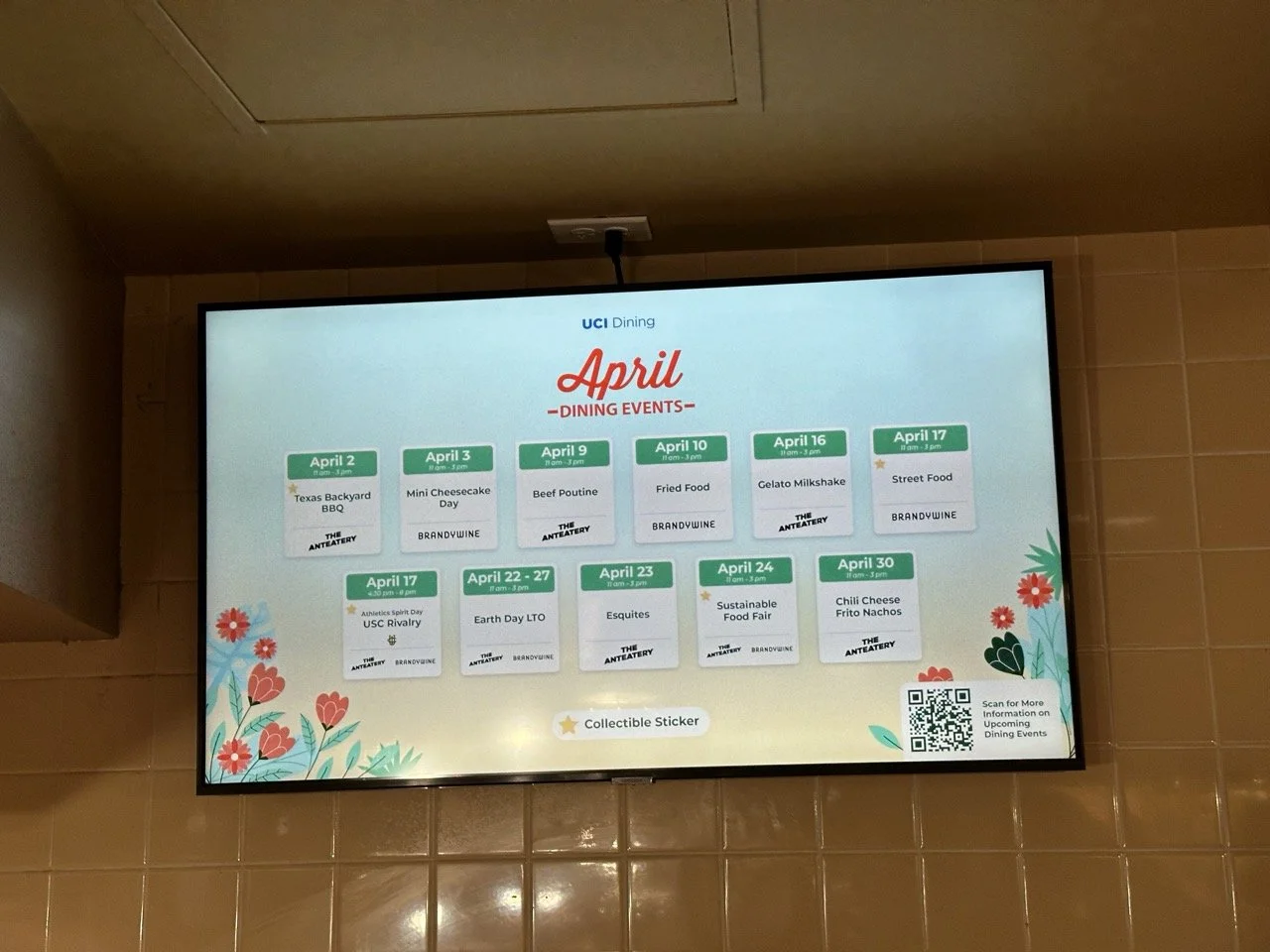 Digital schedule board displaying April dining events at UC Irvine, including BBQ, cheesecake, poutine, fried food, milkshake, street food, and other themed days with dates from April 2 to April 30, decorated with flowers and a QR code for more infor