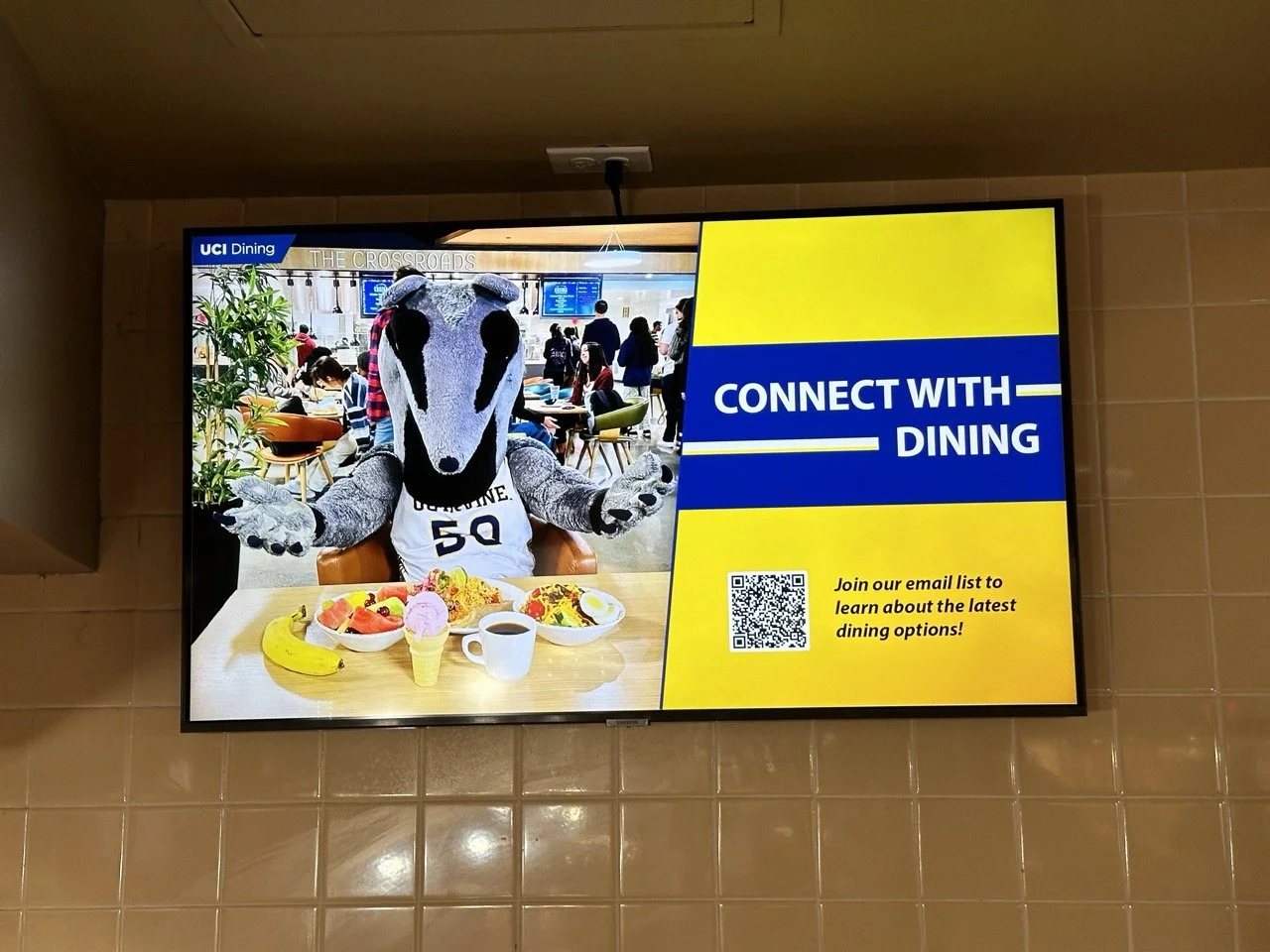 Digital signage promoting connecting with dining, featuring a mascot with a dog head wearing a jersey, sitting at a table with food and drinks in a dining area with people in the background.