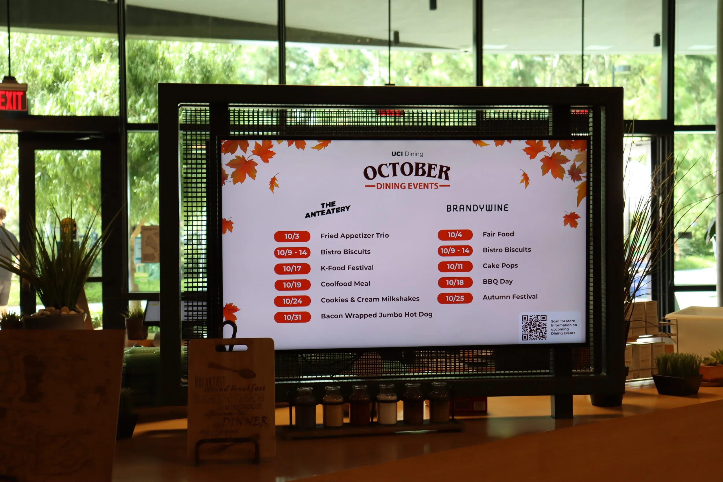 Digital screen displaying October dining events schedule for The Anteater and Brandywine, listing dates and menu items with a QR code in the bottom right corner.