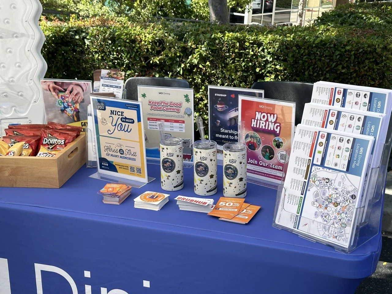 Table display with brochures, flyers, coffee cups, snacks, and recruitment signs outdoors, with a hedge and sidewalk in background.