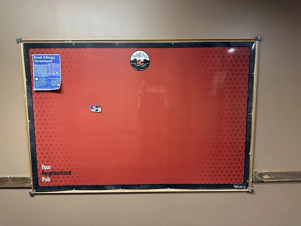Empty red bulletin board with black border and two stickers, one at the top center with a pub logo, and one on the left side noting food allergy awareness, mounted on a tan wall.