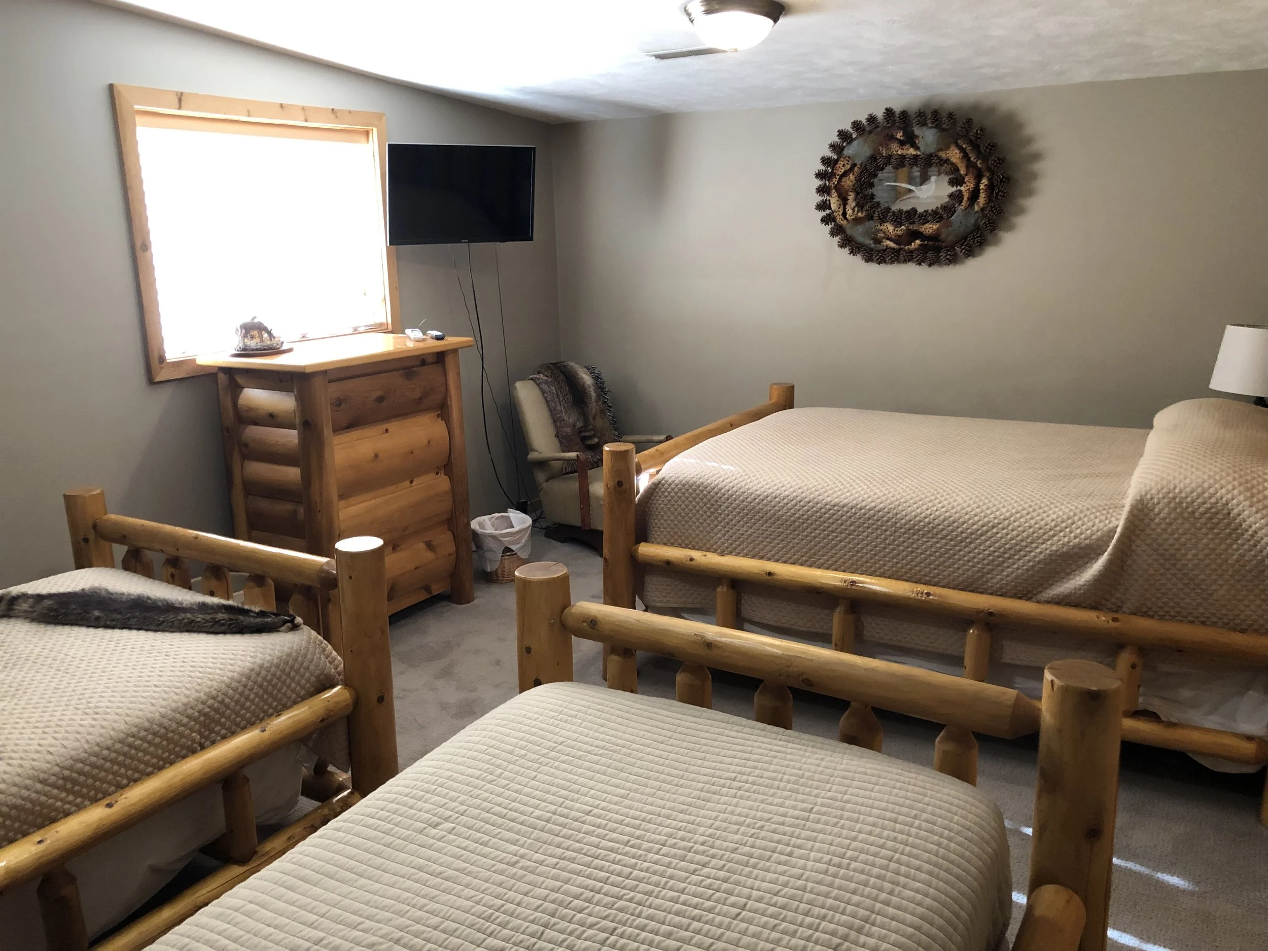 Lodging for our all inclusive Wyoming guided waterfowl hunt with a bedroom with three wooden beds, a window with sunlight, a dresser, a chair with a throw blanket, a mounted TV, a decorative wreath on the wall, and a lamp on a side table.