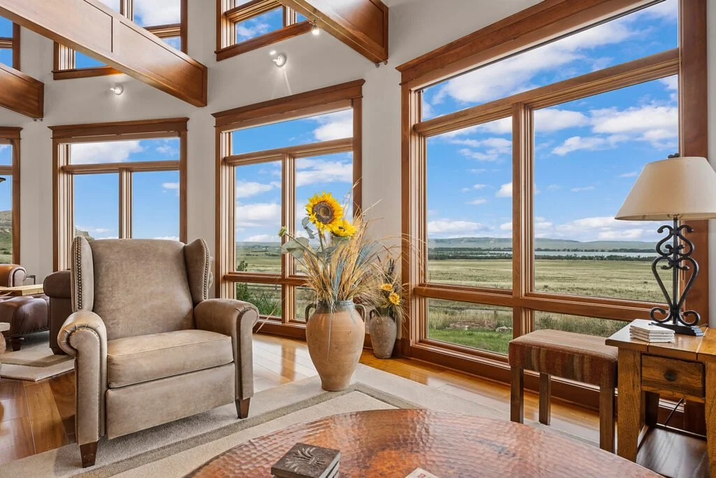 	
Lodging for our all inclusive Wyoming guided waterfowl hunt with a Living room with large windows showcasing a scenic outdoor landscape, beige armchair, wooden side table with a lamp, vase with sunflowers, and hardwood floors.