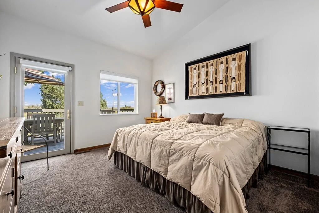 Lodging for our all inclusive Wyoming guided waterfowl hunt with a Bedroom with a large bed, beige bedding, wall art, a nightstand with a lamp, a sliding glass door leading to a balcony, and windows showing outdoor trees and a blue sky.