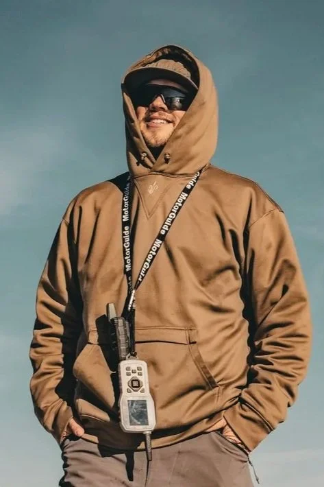 Our professional goose hunting guide who is a young man wearing a brown hoodie, sunglasses, and a cap, smiling against a clear sky. He has a lanyard around his neck with a device attached to it.