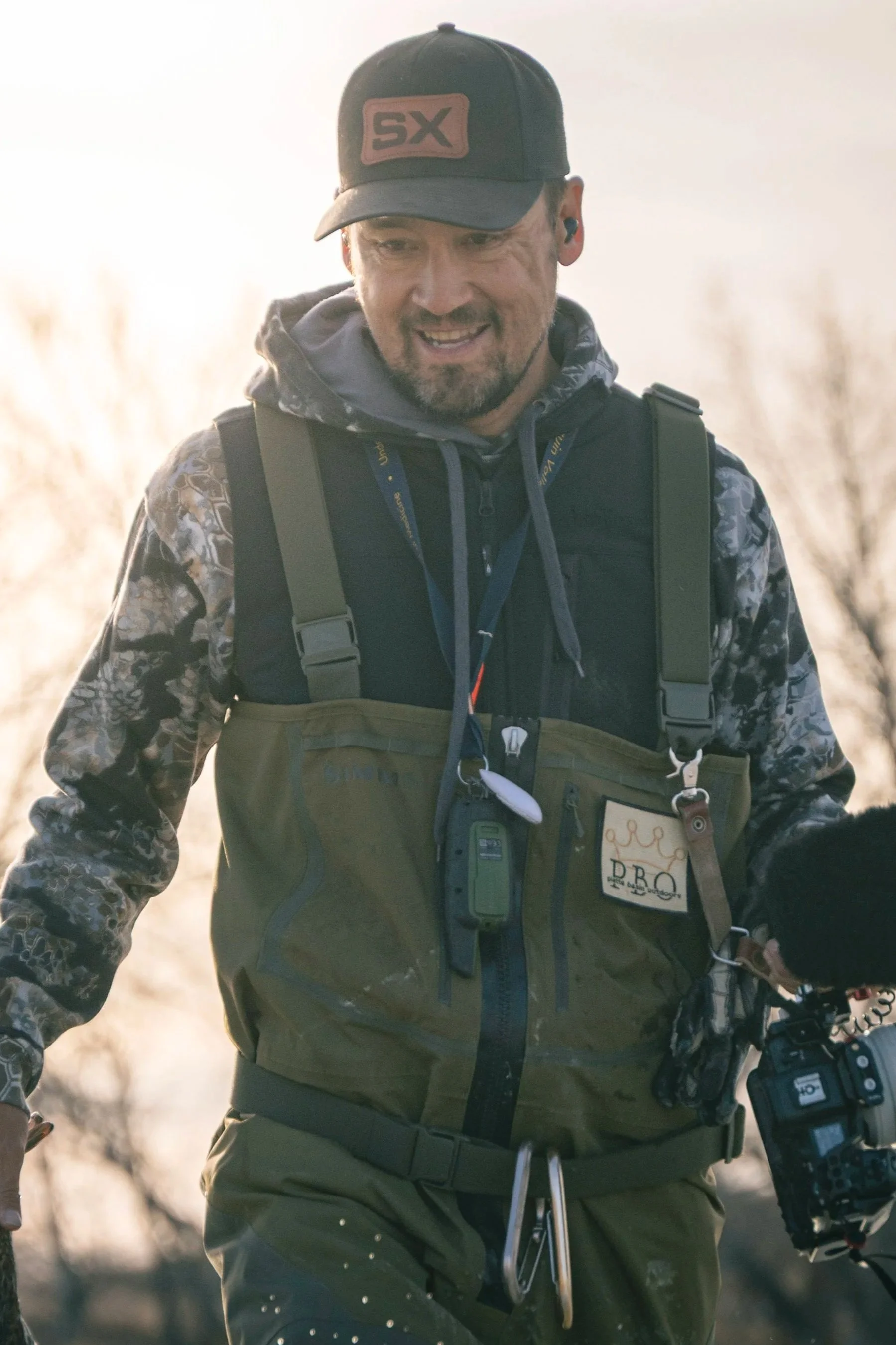 A man with a beard and camouflage cap standing outdoors with four people blurred in the background during sunset.