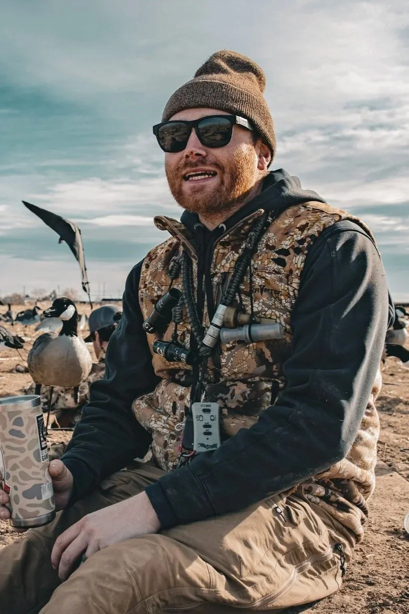 A man in camouflage clothing, wearing sunglasses, a cap, and a backpack, stands outdoors near a body of water with tall grass and bare bushes, under a partly cloudy sky.