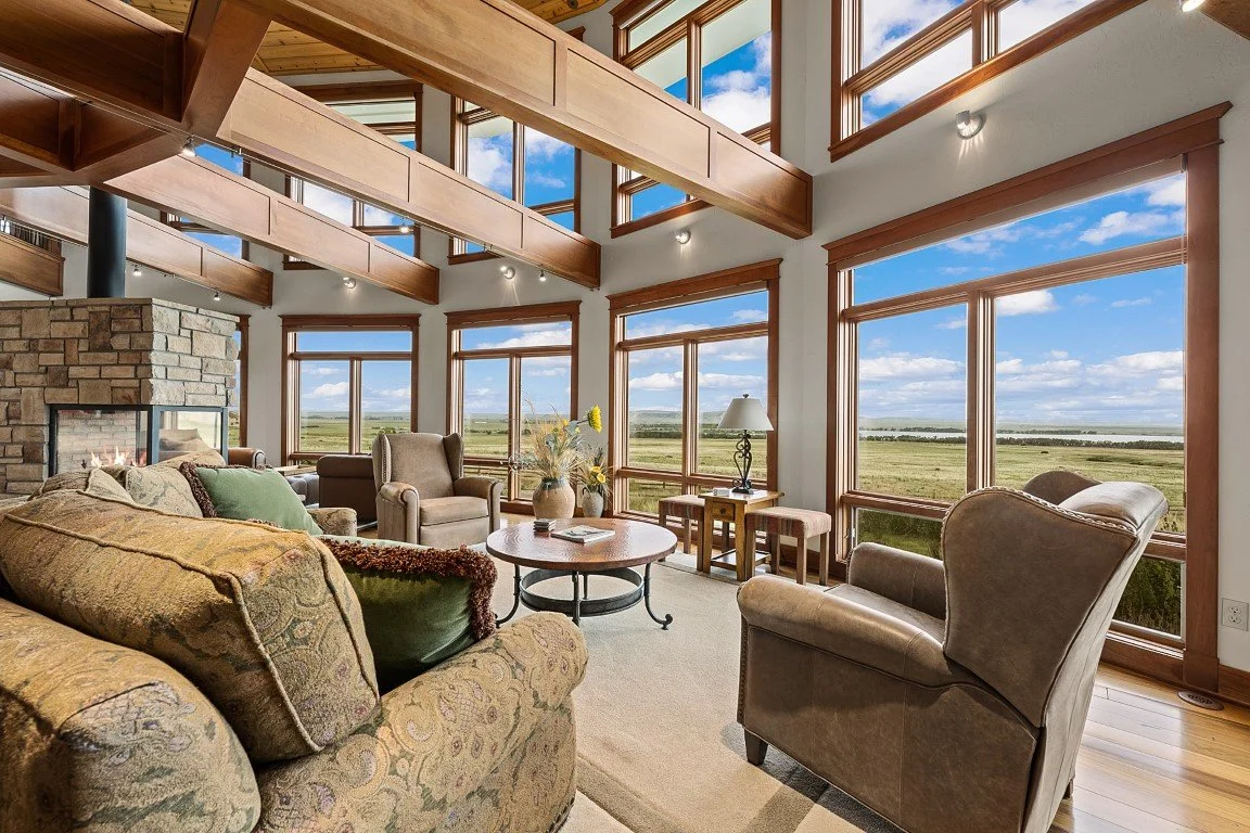 Lodging for our all inclusive Wyoming guided waterfowl hunt with a Spacious living room with large windows showing a scenic outdoor view of fields and a blue sky with clouds, wooden beams, and a stone fireplace.