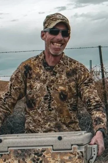 Man wearing camouflage clothing and sunglasses smiling outdoors in front of a chain-link fence.