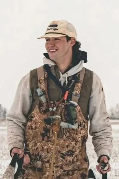 	
Our professional duck hunting guide who is a young man wearing a beige cap, camouflage vest, and outdoor gear, smiling while standing outdoors in a snowy landscape.
