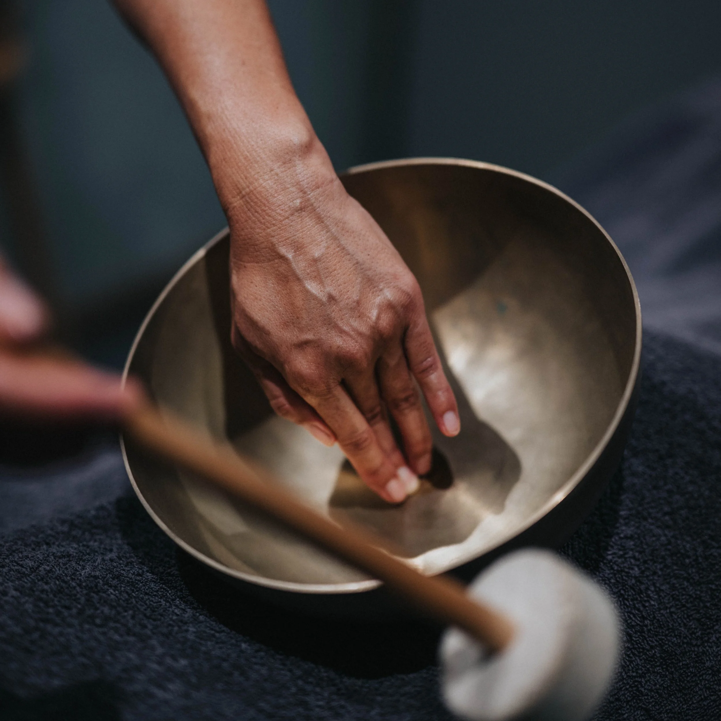 A person’s hand playing a metal singing bowl with a wooden mallet.