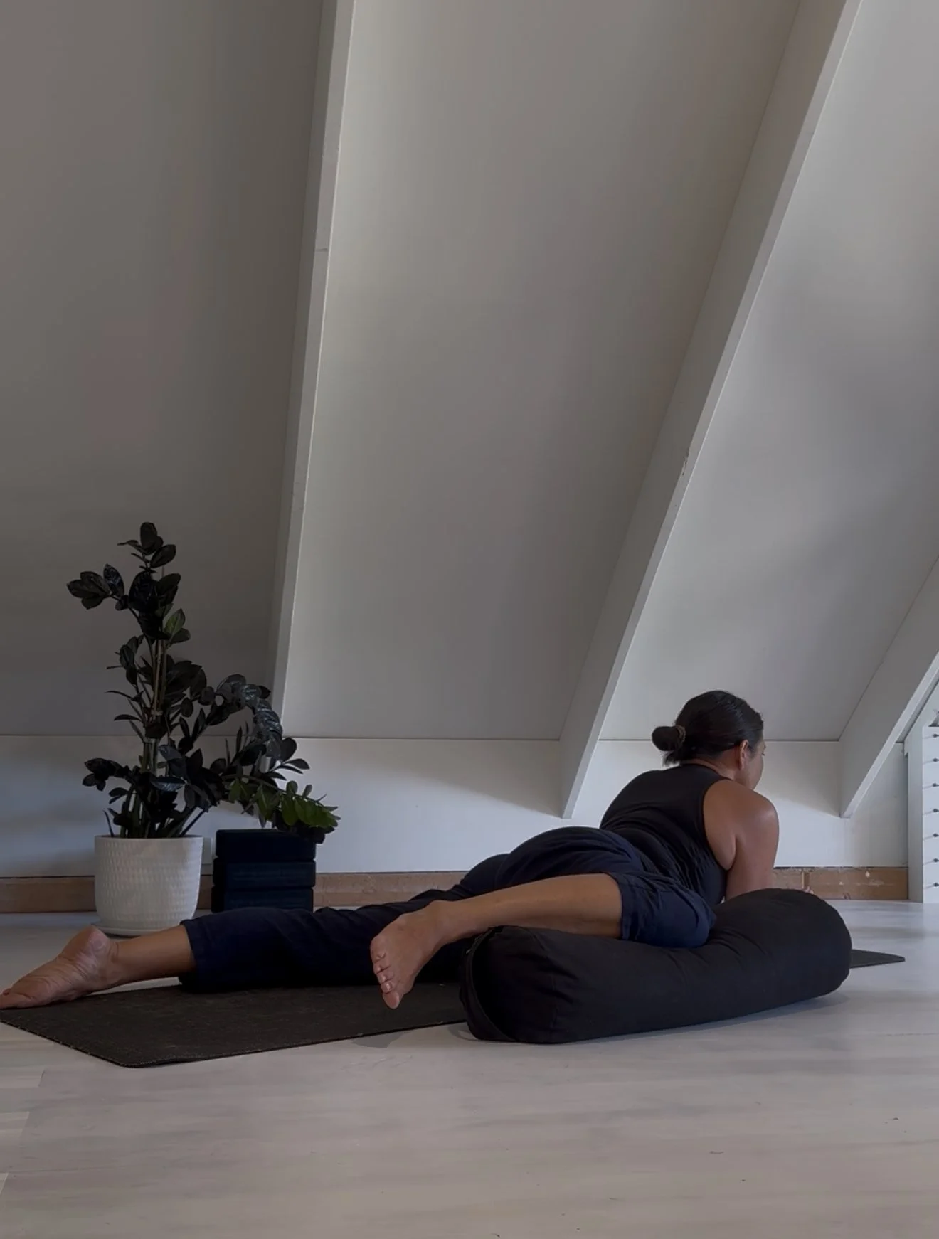A person practicing yoga on a black mat with a cylindrical bolster in a room with a sloped ceiling, next to a potted plant.