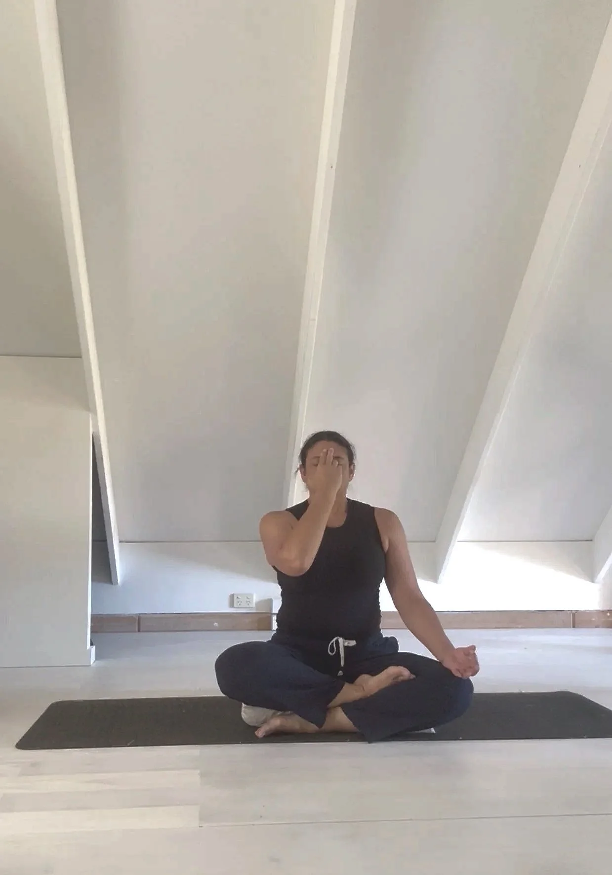 A woman in black workout clothes sitting cross-legged on a yoga mat, practicing meditation or yoga in a room with slanted ceiling and white walls.