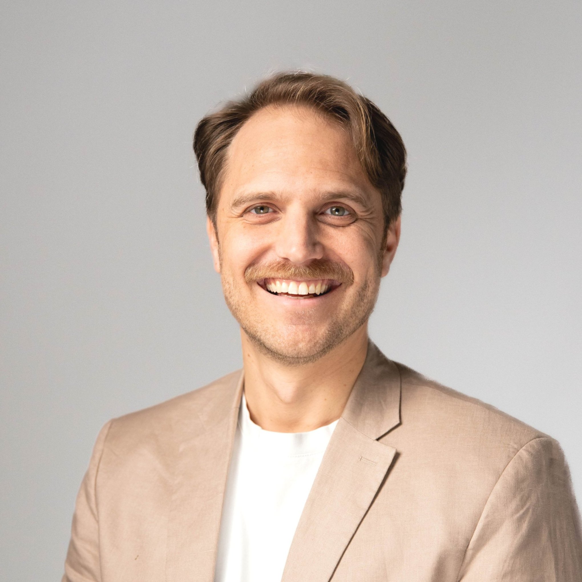 Lächelnder Mann mit beige Jacke vor hellem, neutralem Hintergrund.