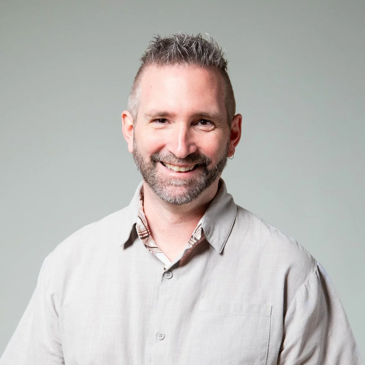 Ein lachender Mann mit kurzgrauem, strukturiertem Haar, Bart und Brille trägt ein hellgraues Hemd vor einem neutralen Hintergrund.