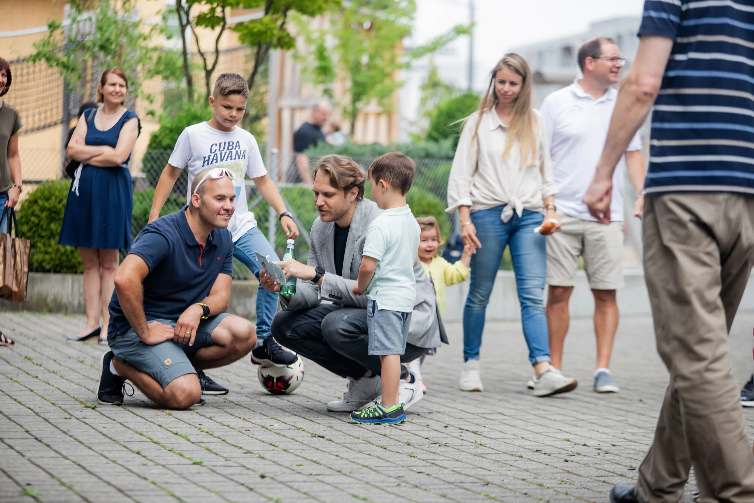 Familie und Freunde auf einem Spielplatz, Kinder mit Ball, Erwachsenen beim Plaudern und lachen, im Freien bei Tageslicht.