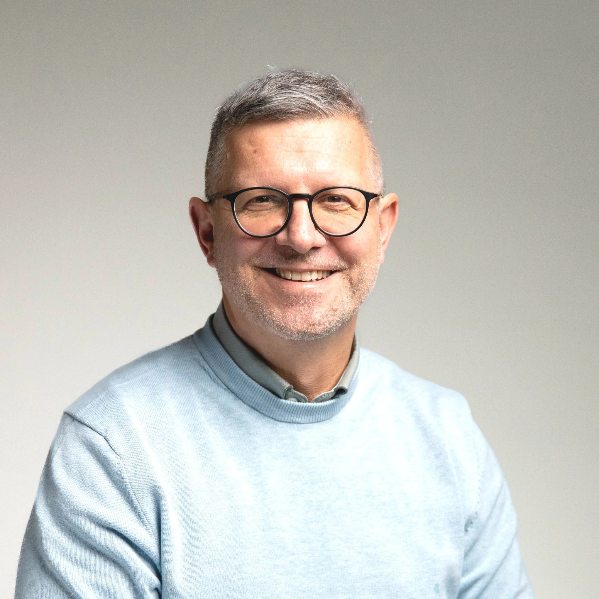 Ein lachender Mann mit Brille, grauem Haar und grauem Bart vor einem neutralen Hintergrund.