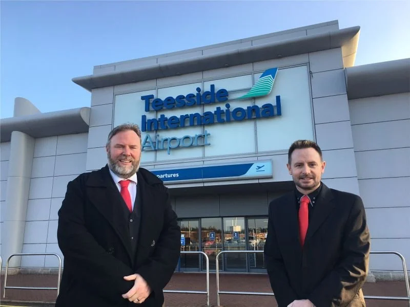 Two smiling men in black coats wearing red ties standing outside Teesside International Airport